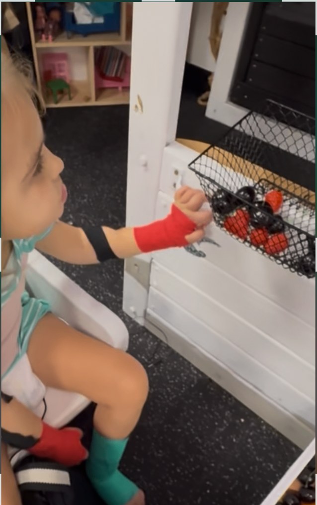 Seated child with the forearm supported at a low shelf, reaching to grasp a small object from a basket at chest height